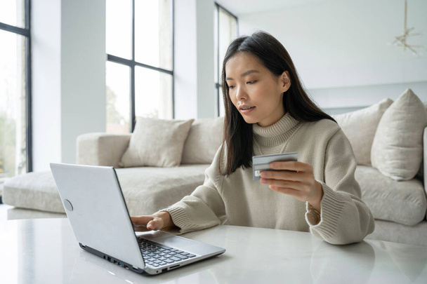 A young Asian woman holding a credit card and using a laptop. A female entrepreneur working from home. Online shopping, the concept of working at home - Zdjęcie, obraz
