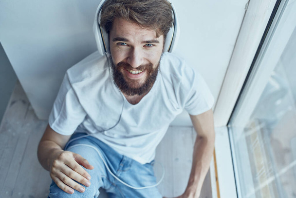 Ο άνθρωπος κάθεται στο περβάζι του παραθύρου με ακουστικά στην επικοινωνία ακουστικών - Φωτογραφία, εικόνα