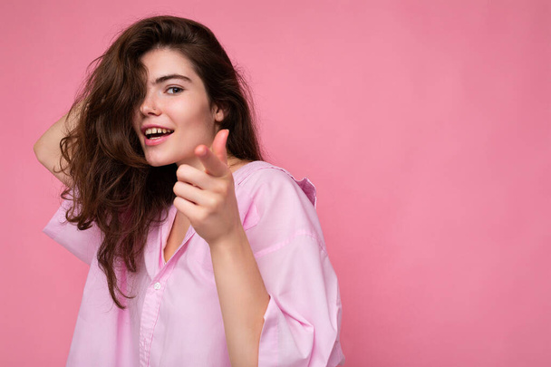 Portret van een mooie vrolijke glimlach positieve jonge krullende brunette vrouw met roze shirt en grijze hoed geïsoleerd op roze achtergrond met kopieerruimte en wijzende vinger naar de camera - Foto, afbeelding