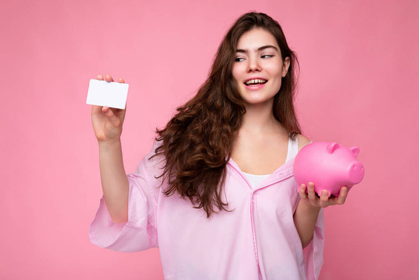 Retrato de hermosa positiva alegre linda sonriente joven morena en camisa elegante aislado sobre fondo rosa con espacio de copia y la celebración de la caja de dinero de cerdo rosa y tarjeta de crédito para maqueta - Foto, imagen
