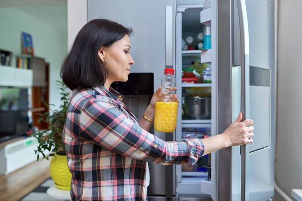 A woman opens the refrigerator at home in the kitchen, holding a bottle of orange juice. - Foto, Imagen