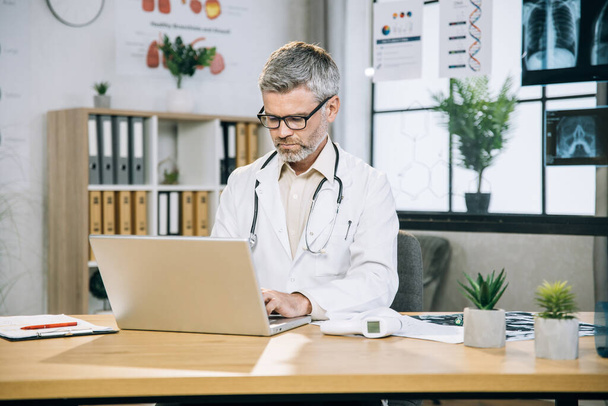 Grey haired doctor sitting at desk and typing on laptop - Photo, image