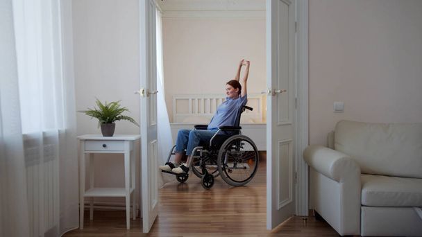 Uma jovem deficiente está andando em sua cadeira de rodas ao redor da sala e alongamento  - Foto, Imagem