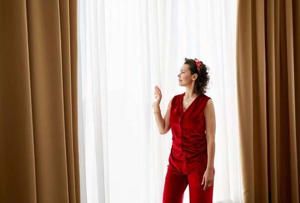 Beautiful young curly woman standing next to the curtains on big panoramic window - 写真・画像