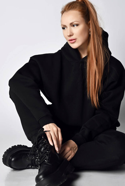 Powerful young sporty woman in black sports suit hoodie, pants and massive brutal army shoes sits on floor looking aside - Fotografie, Obrázek