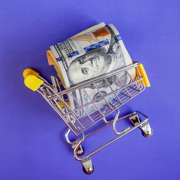 concept - curled dollar banknotes lie in Shopping Cart Trolley against the purple background - Foto, afbeelding