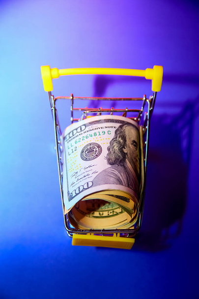 concept - curled dollar banknotes lie in Shopping Cart Trolley against the purple background - Photo, image