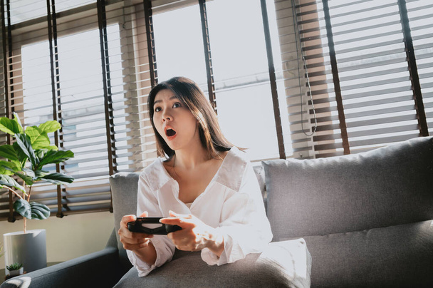Mulher asiática excitada jogando videogame na sala de estar enquanto sentada no sofá em casa - Foto, Imagem