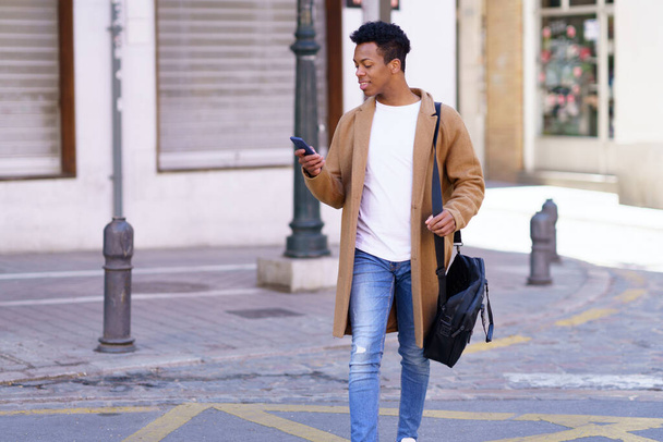 Νεαρός μαύρος κοιτάζει το smartphone του ενώ περπατάει στο δρόμο. - Φωτογραφία, εικόνα