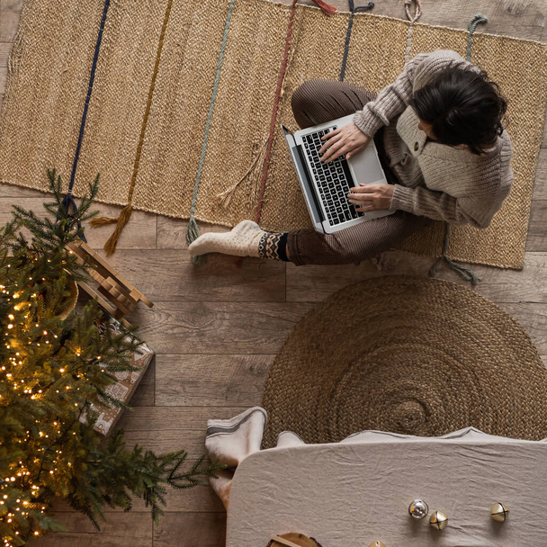 Esteettinen ylhäältä näkymä nainen istuu matolla ja työskennellä kannettavan tietokoneen kodikas mukava koti olohuone sisustettu jouluksi, uudenvuoden loma juhla. Joulukuusi, hehkuvat valot - Valokuva, kuva