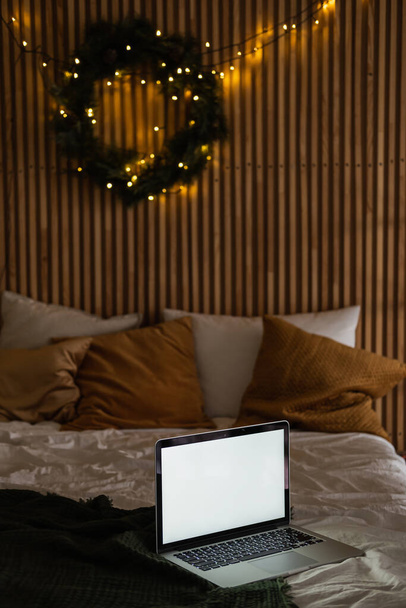 Laptop computer in bed. Christmas wreath, warm garland light on wooden wall. Aesthetic morning concept. Freelancer, work at home business layout. - Photo, image