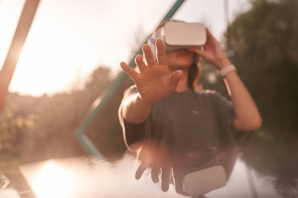 Ένα κορίτσι με γυαλιά εικονικής πραγματικότητας στο κεφάλι με τα χέρια της. Απλώνει το ένα χέρι στην κάμερα, δίπλα σε μια ακτίνα φωτός.. - Φωτογραφία, εικόνα