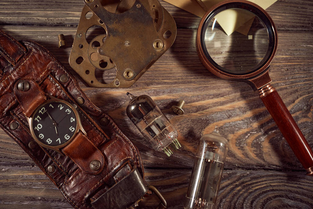 スチームパンクまだ生活-木製の背景に古いヴィンテージオブジェクト.レザー手首腕時計、ダークグラス、古いチューブランプ、虫眼鏡、ギア - 写真・画像