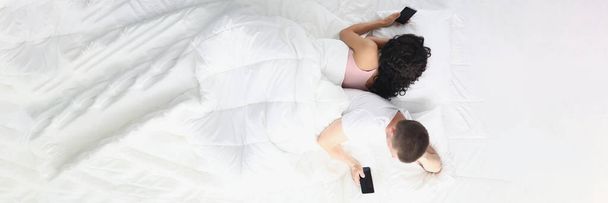 Uomo e donna sdraiati in camera da letto sul letto tenendo gli smartphone in mano e allontanandosi l'uno dall'altro - Foto, immagini