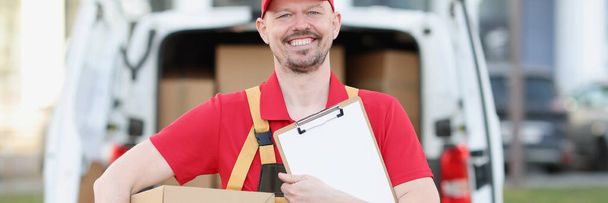 Mensageiro masculino em uniforme detém prancheta e caixa de papelão no fundo do caminhão com caixas - Foto, Imagem