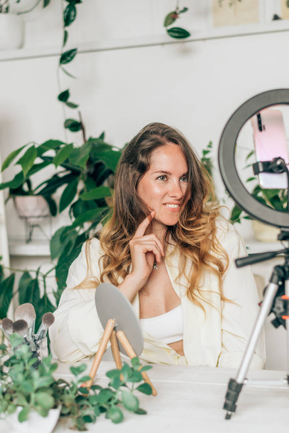 A young woman beauty blogger broadcasts live streaming to review makeup and cosmetic products on social media,reviews beauty product for video blog.Influencer uses smartphone on tripod and ring lamp. - Fotografie, Obrázek