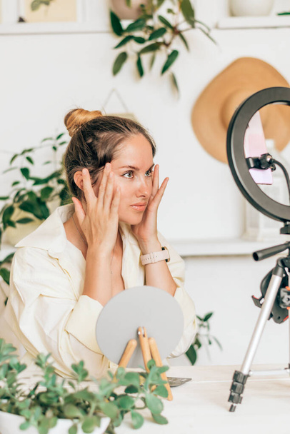 A young woman beauty blogger broadcasts live streaming to review makeup and cosmetic products on social media,reviews beauty product for video blog.Influencer uses smartphone on tripod and ring lamp. - Фото, зображення