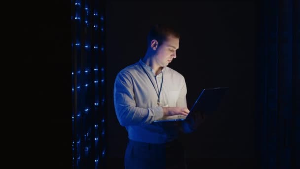 Young man holds device in hand and looks at screen, inspects equipment or hardware rack. Male programmer working with laptop and supporting service while standing in data center spbas. - Footage, Video