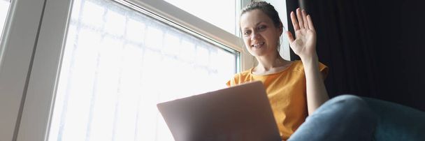 Young woman waving welcome at laptop while sitting on windowsill - 写真・画像
