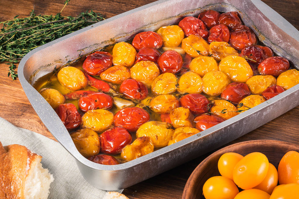 Cooked baked cherry tomatoes with garlic, thyme and pepper with olive oil in a baking sheet - making French homemade tomato confit, close-up - Photo, Image