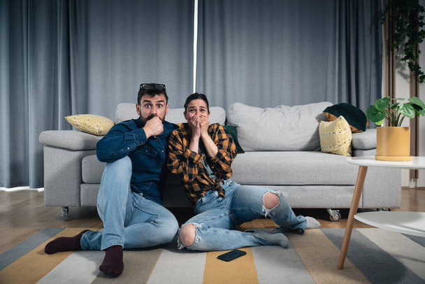 Young couple sitting on the floor of their apartment watching scary horror movie while woman is very scared and man hugs her and she cover her scared face with her hands  - Valokuva, kuva