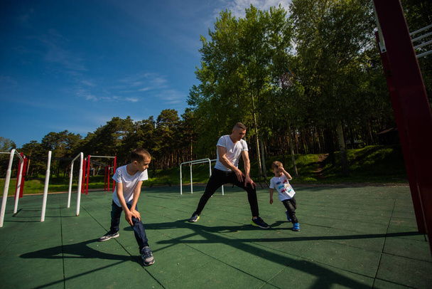 Caucasian man and two boys doing exercises outdoors. Father and sons train on the sports ground - Zdjęcie, obraz