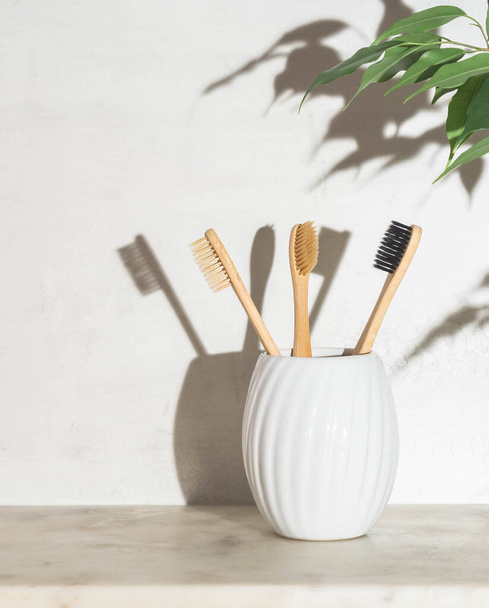 Eco bamboo toothbrushes in ceramic glass on marble shelf and houseplant leaves in the bathroom. Front view. Zero weste care concept - Photo, Image