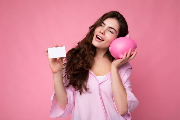 Atractiva joven morena feliz vistiendo camisa aislada sobre fondo rosa con espacio vacío y sosteniendo caja de dinero de cerdo rosa y tarjeta de crédito para maqueta. Concepto bancario - Foto, imagen