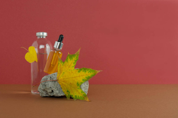 Tropfflasche mit Gelserum und Vogelbeeren auf rotem Hintergrund, einem Stein und einer Flasche mit Flüssigkeit. Herbstblätter. - Foto, Bild
