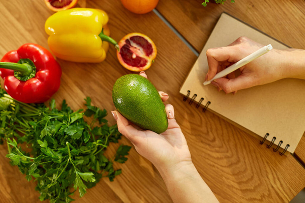 Γυναίκα σχεδιάζει, γράφοντας εβδομαδιαία γεύματα σε ένα σημείωμα σχεδιαστή γευμάτων ή σχέδιο διατροφής σε ξύλινο τραπέζι με υγιεινά φρούτα και λαχανικά τροφίμων στην κουζίνα της στο σπίτι. - Φωτογραφία, εικόνα