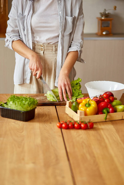 Woman is preparing vegetable salad in the kitchen, cutting leaf of salad on wooden cutting board, Healthy food concept, vegan or diet. - Photo, image
