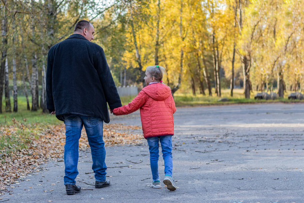 un joven padre encorvado en ropa negra, cojeando en una pierna, camina por el parque de otoño con su hija. Primer plano. espacio de copia, - Foto, imagen