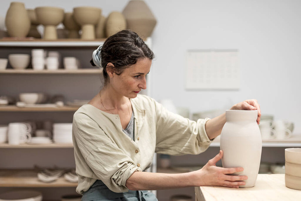 Young woman holding pottery instrument for scraping, smoothing, shaping and sculpting. Lady siting on bench with pottery wheel and making clay pot - Foto, Bild