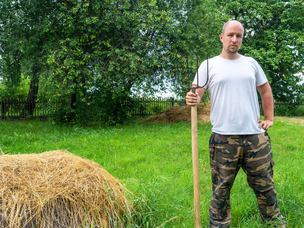 Мужчина стоит спиной к камере с вилами перед стогом сена. Сельский пейзаж, подготовка корма для животных. Сельская жизнь - Фото, изображение