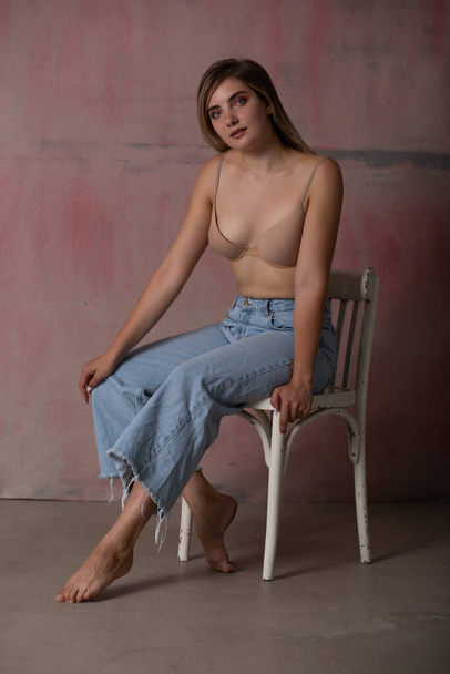 Gorgeous blonde in a bra and jeans sitting on a chair - Фото, зображення
