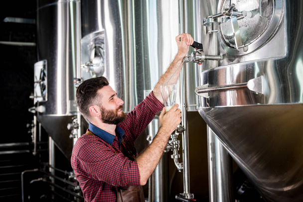 革のエプロンを着た若い醸造者が現代の醸造所でビールをテストしています - 写真・画像