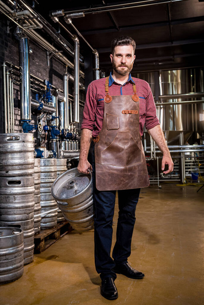 Young male brewer in leather apron holds barrel with craft beer at modern brewery factory - 写真・画像