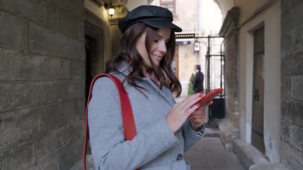  Das junge hübsche Mädchen im Herbstkleid mit Zahnspange lächelt aufrichtig und benutzt ein Smartphone. Schöne Frau spaziert mit Smartphone in der Hand durch die Altstadt  - Filmmaterial, Video