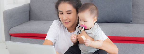 Młoda azjatycka matka pracuje na laptopie, a córka opieki w domu, mama i dziewczynka siedzi za pomocą notebooka wideo rozmowy, rodzica i więzi, w pomieszczeniach, rodziny i koncepcji biznesu. - Zdjęcie, obraz