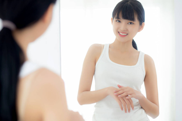 Krásná mladá asijská žena hledá zrcadlo a použití hydratační krém a krém s rukama pro ochranu pokožky, péče o pleť a kosmetické pro krásu, ošetření a péči o tělo, péče o pleť. - Fotografie, Obrázek