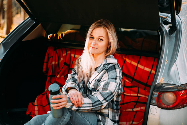 a blonde adult girl in a plaid shirt is drinking coffee sitting in the open trunk of a car. picnic in the car. - Φωτογραφία, εικόνα