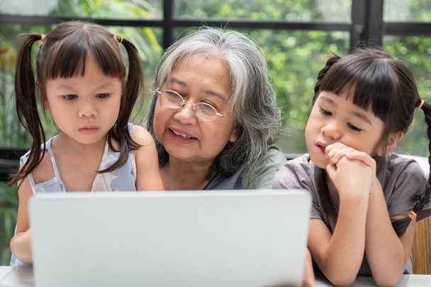 Ασιατική γιαγιά με τα δύο εγγόνια της να διασκεδάζουν και να παίζουν online εκπαιδευτικά παιχνίδια με ένα σημειωματάριο υπολογιστή στο σπίτι στο σαλόνι. Έννοια της online εκπαίδευσης και φροντίδας από τους γονείς. - Φωτογραφία, εικόνα