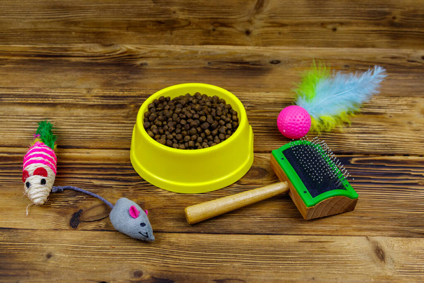 Comida seca para gatos en tazón, juguetes para gatos y cepillo de mimbre para mascotas sobre fondo de madera. concepto de cuidado de mascotas - Foto, imagen