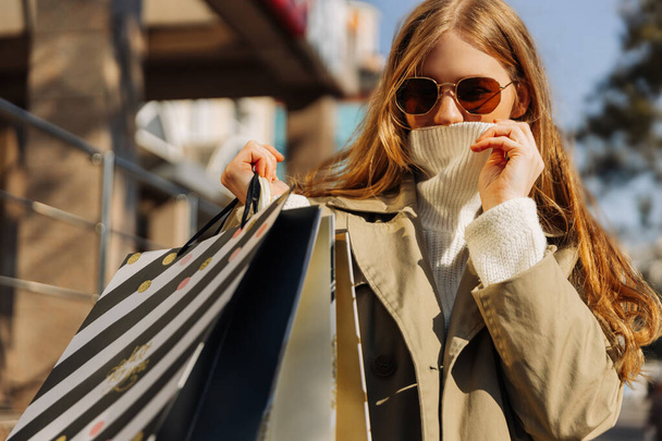 hermosa mujer en impermeable beige con gafas de sol y bolsas de compras en un suéter blanco, cubre su cara con luz del frío. otoño tiempo, venta, compras al aire libre - Foto, imagen