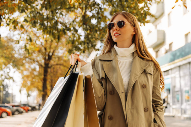 stylish woman in a beige raincoat and sun goggles with shopping bags in her hands, on against the autumn trees, season of discounts, sale. business woman shopping in city - Photo, Image