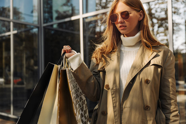 blonde european woman in sunglasses in a beige raincoat and with shopping bags in her hands stands in front of a glass building, a shopping center, shopping in the city, outdoors - Foto, afbeelding