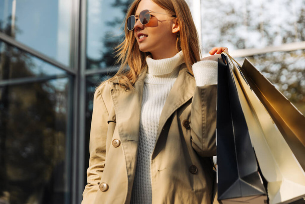 blonde european woman in sunglasses in a beige raincoat and with shopping bags in her hands stands in front of a glass building, a shopping center, shopping in the city, outdoors - Photo, Image
