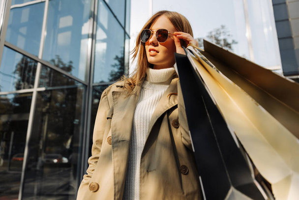 blonde european woman in sunglasses in a beige raincoat and with shopping bags in her hands stands in front of a glass building, a shopping center, shopping in the city, outdoors - Photo, Image