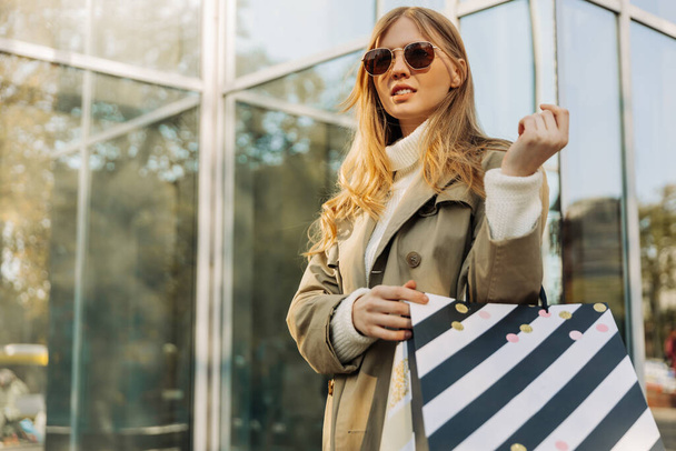 Μια όμορφη ξανθιά με γυαλιά ηλίου και τσάντες για ψώνια στέκεται πάνω σε ένα γυάλινο κτίριο, με ένα μπεζ αδιάβροχο. concept: φθινόπωρο ψώνια, μαύρη Παρασκευή, Νοέμβριος - Φωτογραφία, εικόνα