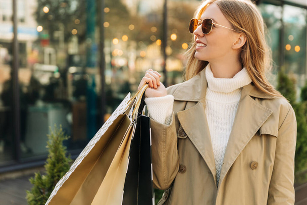 a beautiful woman in the sun of goggles in a beige raincoat and shopping bags in her hands, stands in front of a glass building, and lights. autumn theme: shopping. - Photo, image